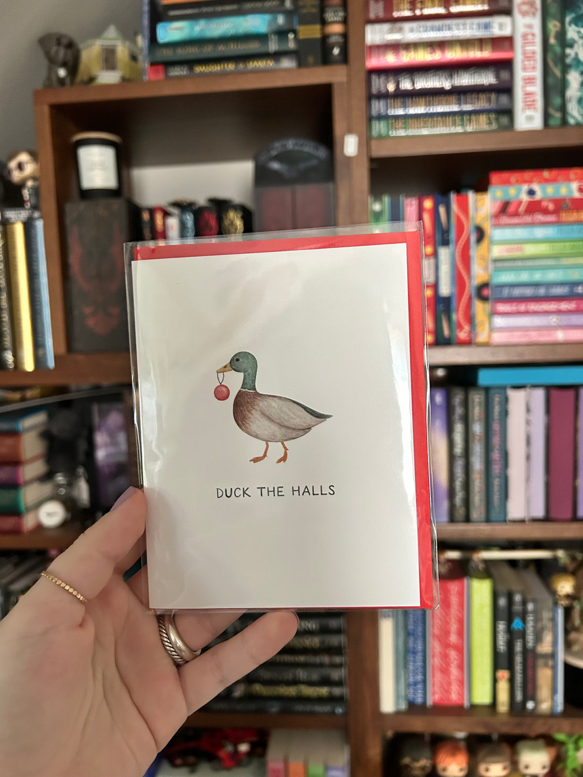 A hand holding a white greeting card with a red envelope and an illustration of a mallard duck holding a red apple, with the text 'DUCK THE HALLS', in front of a bookshelf.