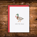 A white greeting card with a red envelope, featuring a watercolor illustration of a mallard duck holding a red ornament and the text 'DUCK THE HALLS' below, displayed on a rustic wooden background.
