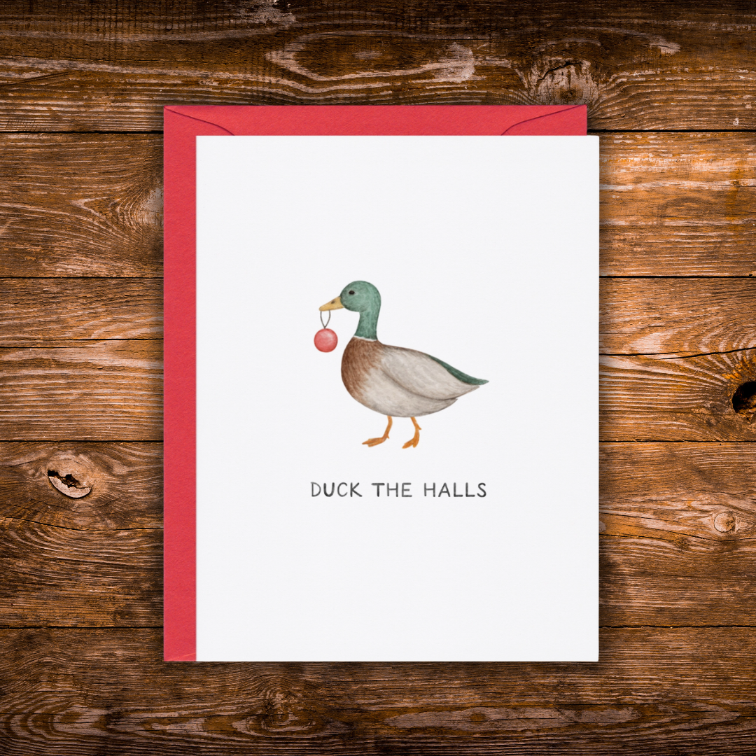 A white greeting card with a red envelope, featuring a watercolor illustration of a mallard duck holding a red ornament and the text 'DUCK THE HALLS' below, displayed on a rustic wooden background.