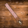 A fabric wristlet strap with a pink and white floral pattern, brown leather trim, and a gold metal clasp, laid diagonally on a rustic wooden plank background.