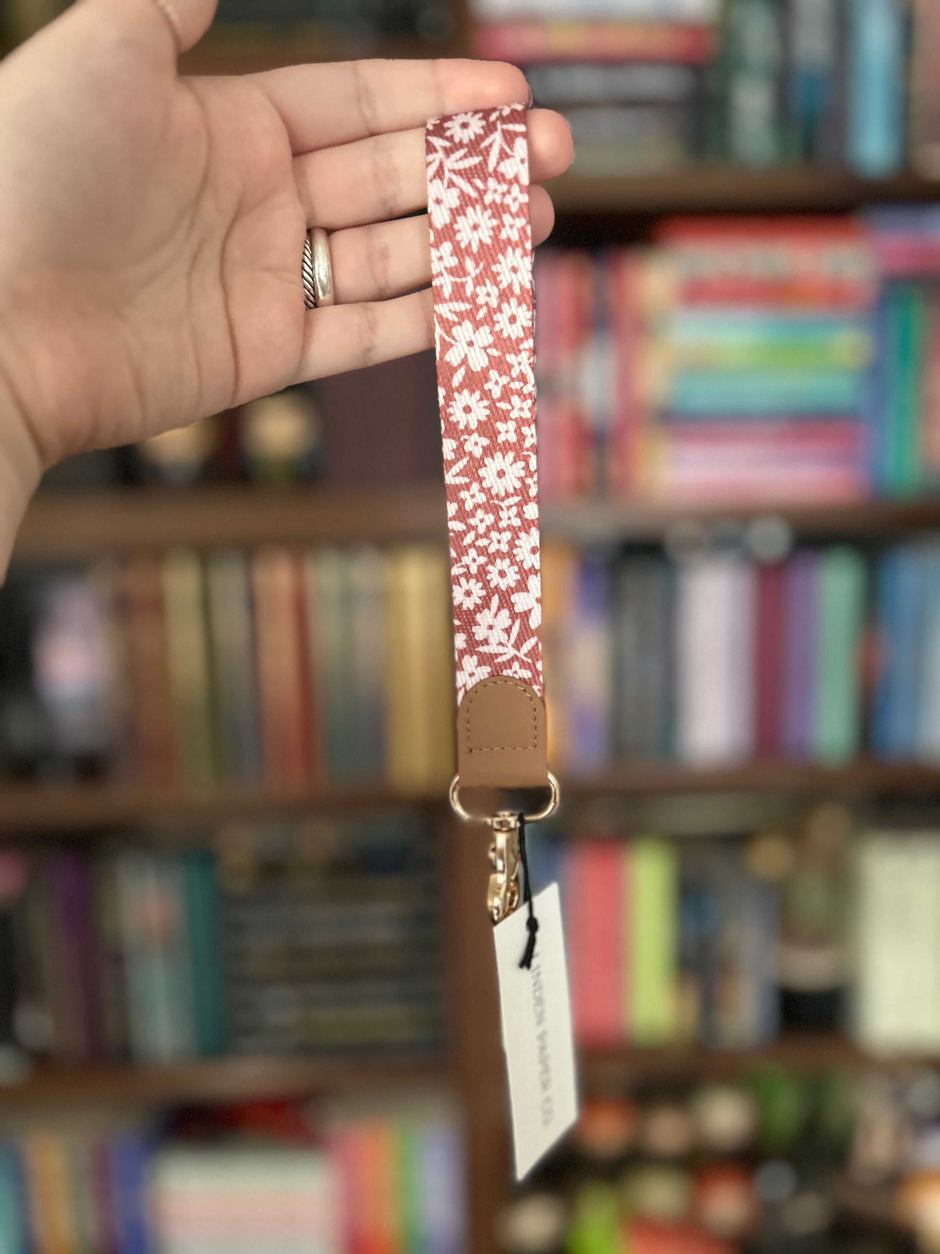 A hand holds a dusty pink floral patterned fabric wrist strap with a brown leather tab and gold-tone metal hardware, set against a blurred background of bookshelves.