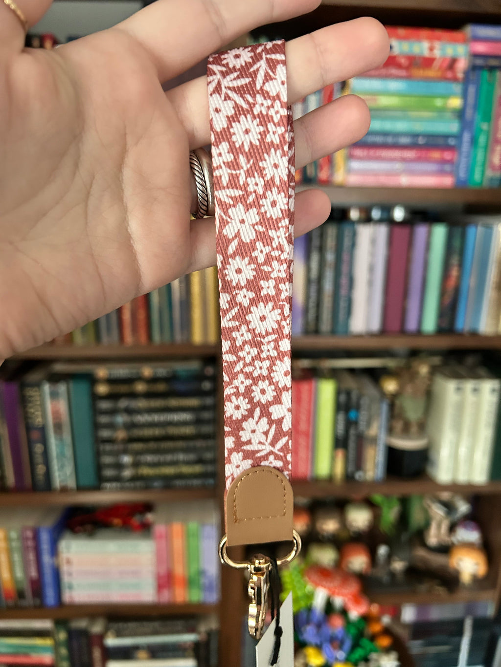 A close-up of a hand holding a fabric wristlet strap with a white floral pattern on a dusty rose background and a tan leather tab, with a blurred bookshelf in the background.