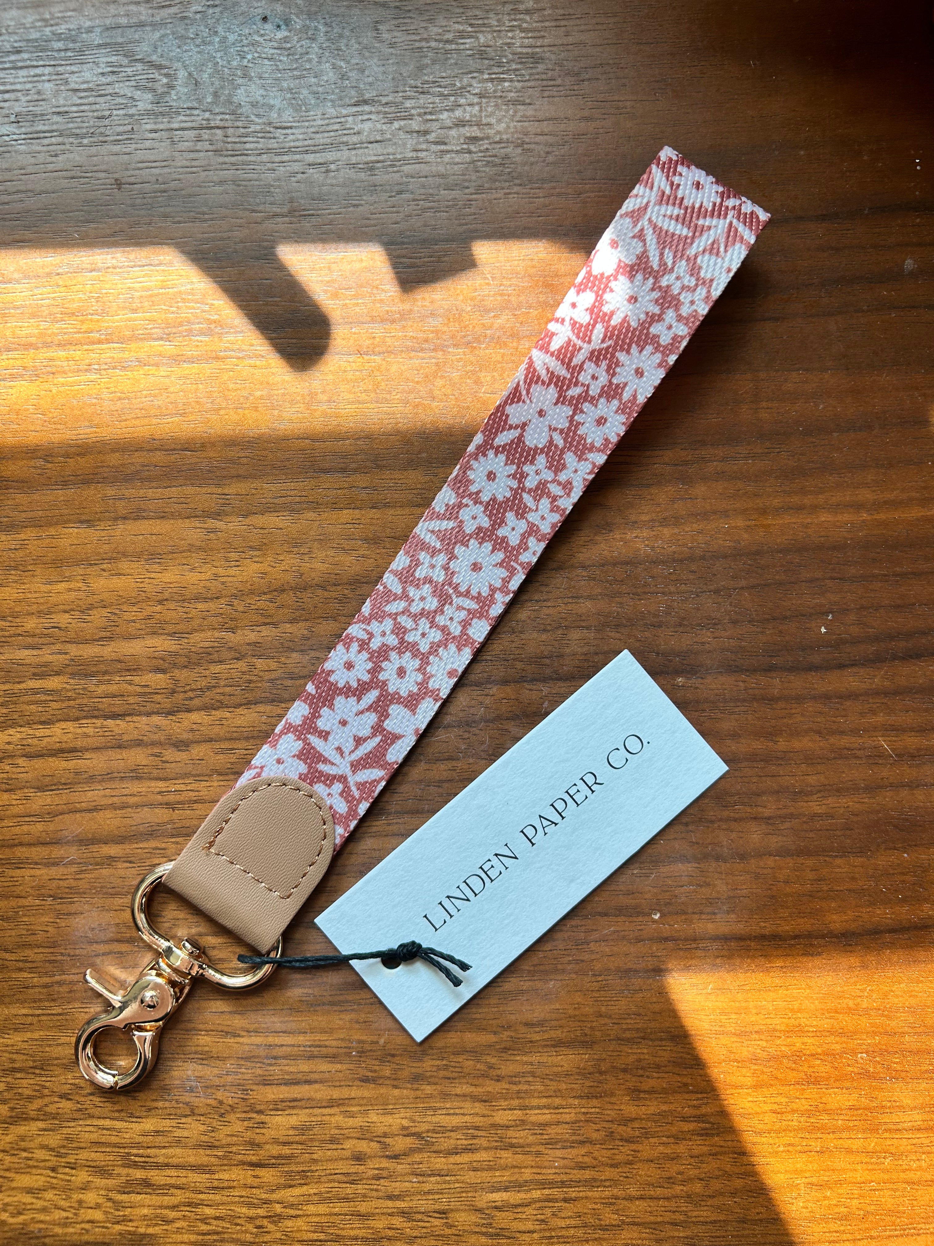 A fabric wristlet strap with a white floral pattern on a dusty rose background, finished with tan leather trim and a gold-tone lobster clasp. A white paper tag reading 'LINDEN PAPER CO.