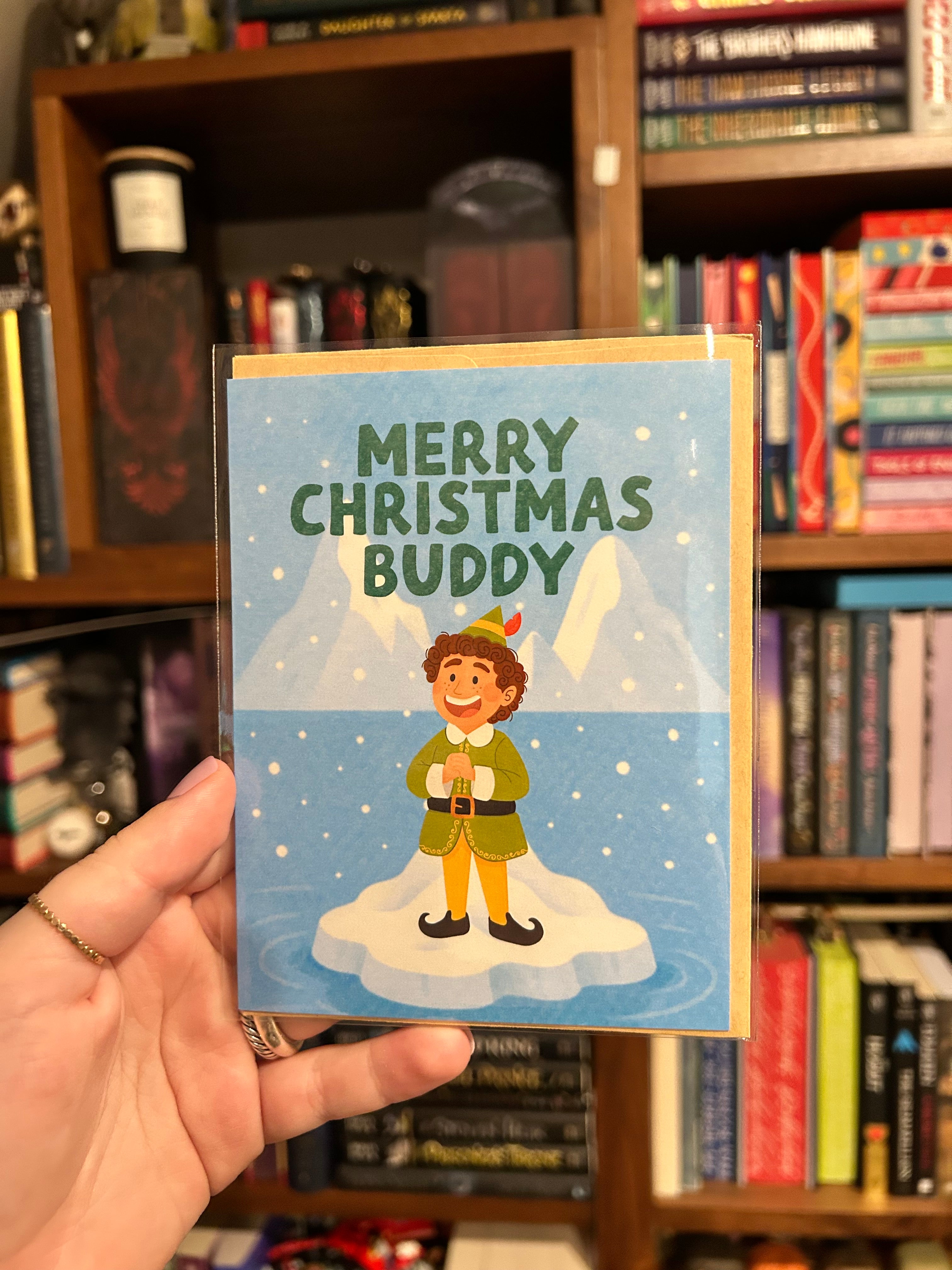 A hand holds a Christmas greeting card in front of a blurred bookshelf. The card features the text 'MERRY CHRISTMAS BUDDY' above an illustration of a person in an elf costume standing on a snowy platform.