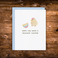 Cute Easter greeting card featuring a fluffy baby chick wearing an eggshell hat beside a cracked pastel Easter egg with the message "Hope You Have a Crackin' Easter."