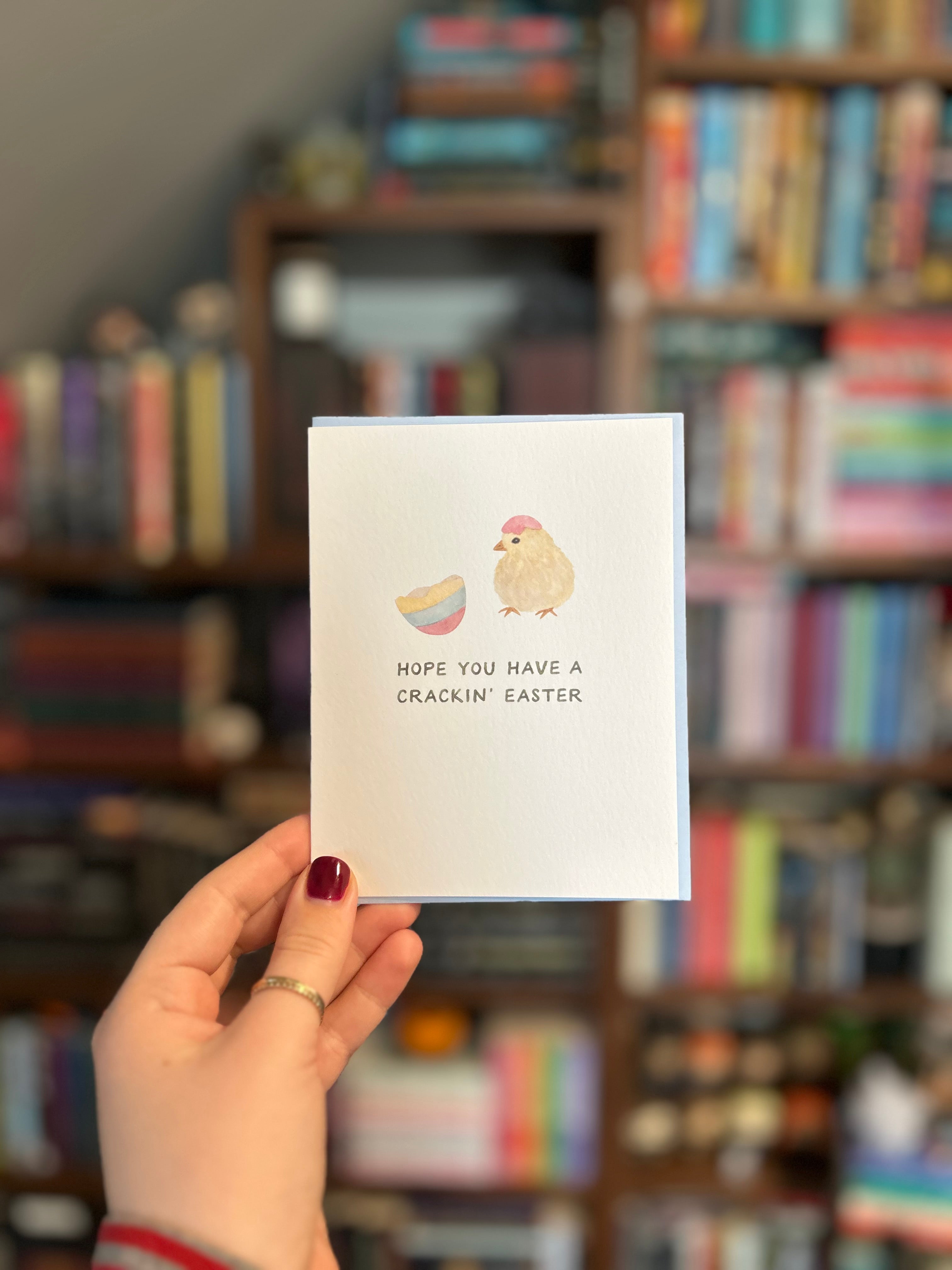 Cute Easter greeting card featuring a fluffy baby chick wearing an eggshell hat beside a cracked pastel Easter egg with the message "Hope You Have a Crackin' Easter."