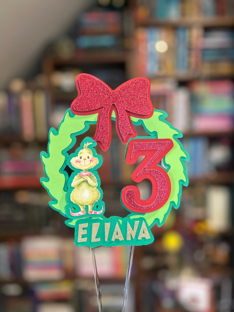 A personalized Christmas cake topper featuring a green glitter wreath, a red glitter bow, a Grinch figure, a large red number 3, and the name 'ELIANA' on a teal banner, shown with metal wire stakes against a blurred bookshelf background.