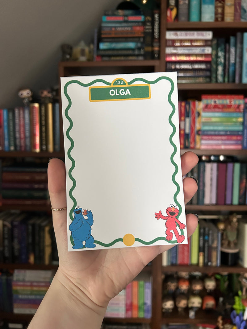 A hand holds a white notepad featuring a green wavy border, a green header that reads 'OLGA', and Sesame Street cartoon character illustrations at the bottom corners, with a blurred bookshelf in the background.