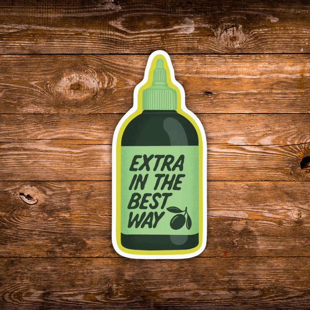 A die-cut sticker shaped like a squeeze bottle with the text 'EXTRA IN THE BEST WAY' and an olive branch graphic, shown on a rustic brown wooden background.