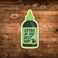 A die-cut sticker shaped like a squeeze bottle with the text 'EXTRA IN THE BEST WAY' and an olive branch graphic, shown on a rustic brown wooden background.