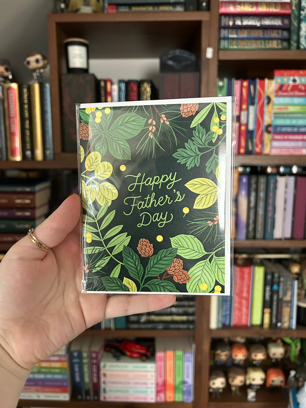 Father's Day greeting card featuring lush green botanical foliage and pinecones surrounding the words "Happy Father's Day" on a dark forest green background.