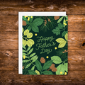 Father's Day greeting card featuring lush green botanical foliage and pinecones surrounding the words "Happy Father's Day" on a dark forest green background.