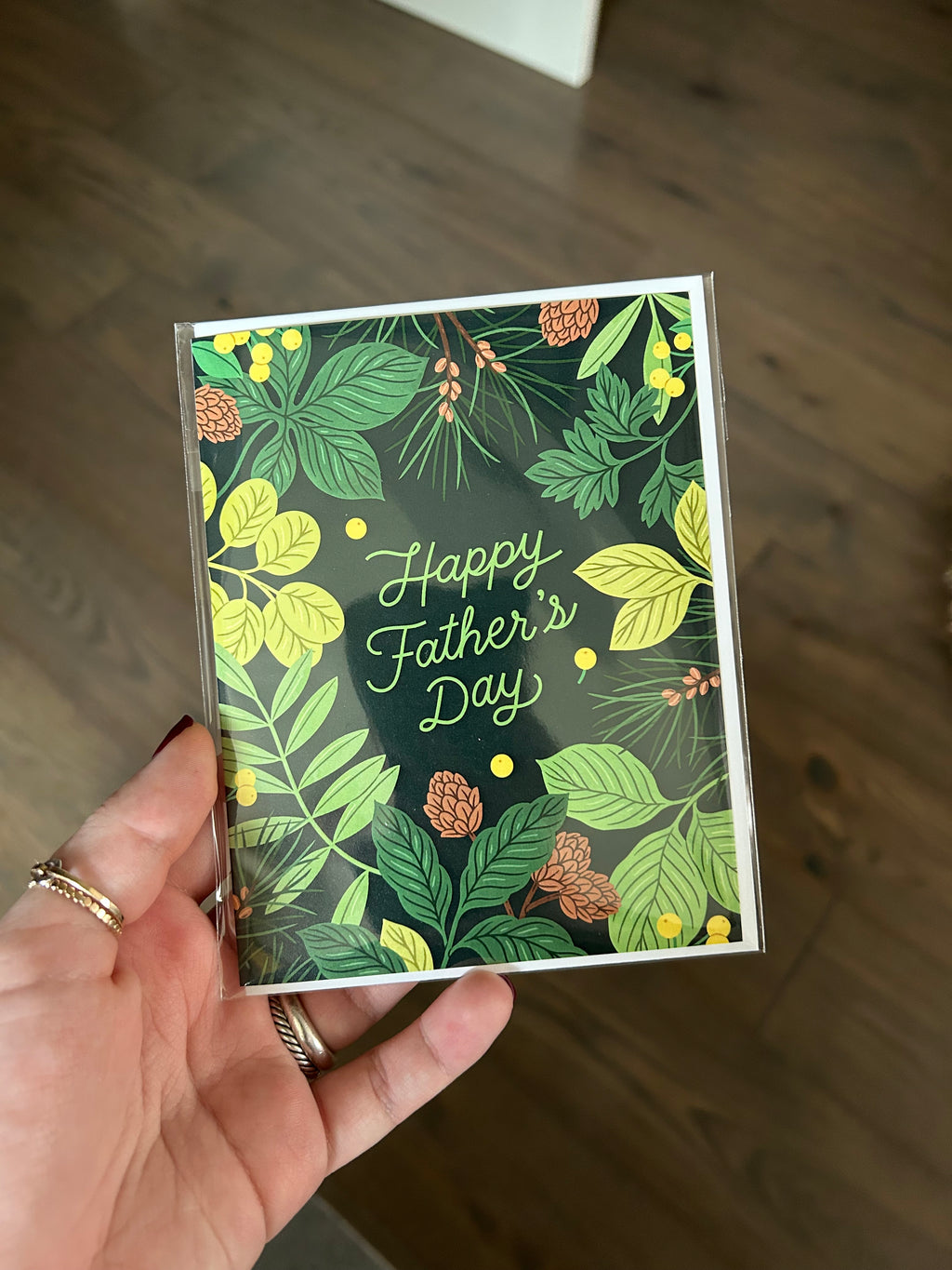Father's Day greeting card featuring lush green botanical foliage and pinecones surrounding the words "Happy Father's Day" on a dark forest green background.
