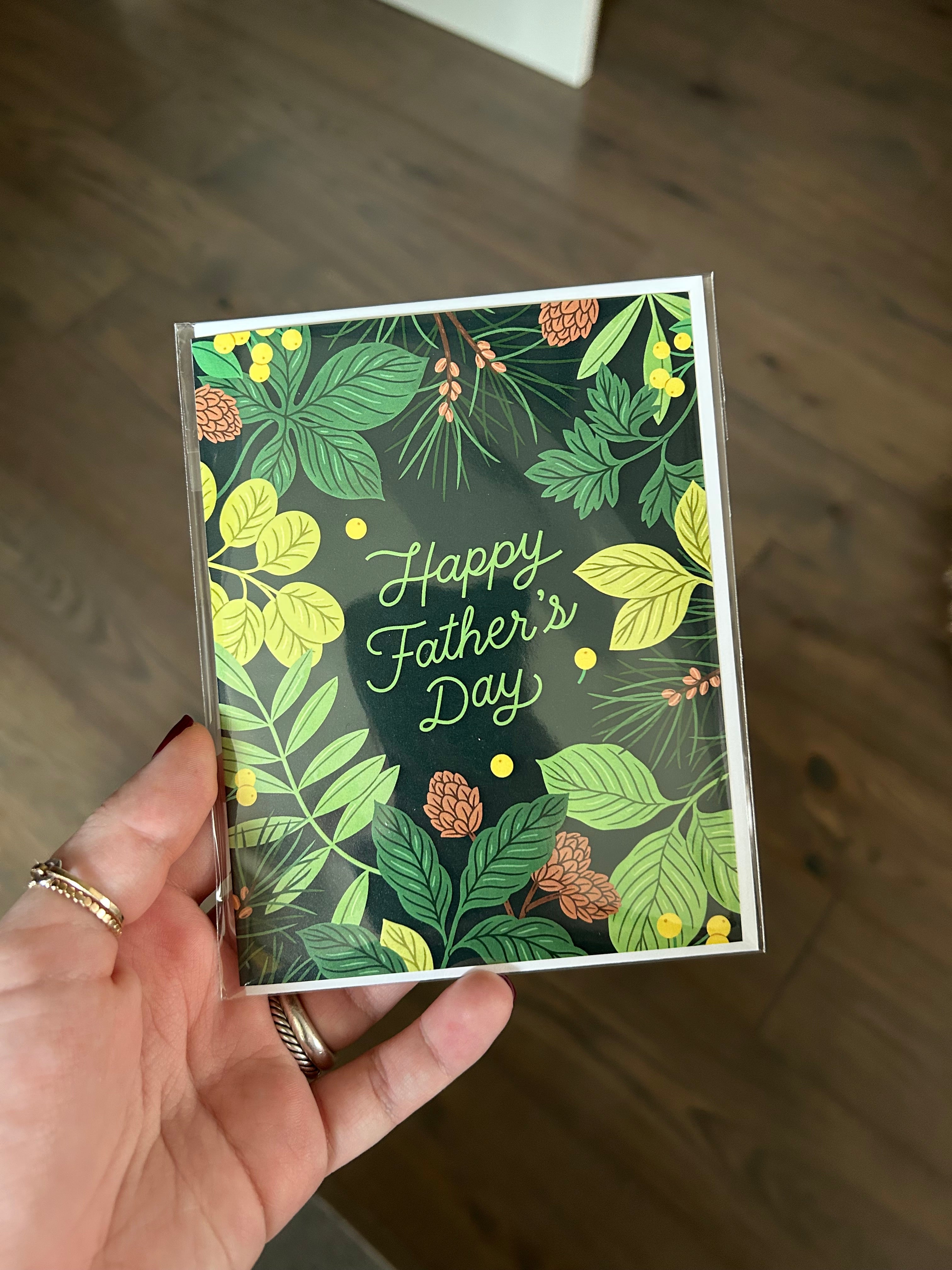Father's Day greeting card featuring lush green botanical foliage and pinecones surrounding the words "Happy Father's Day" on a dark forest green background.