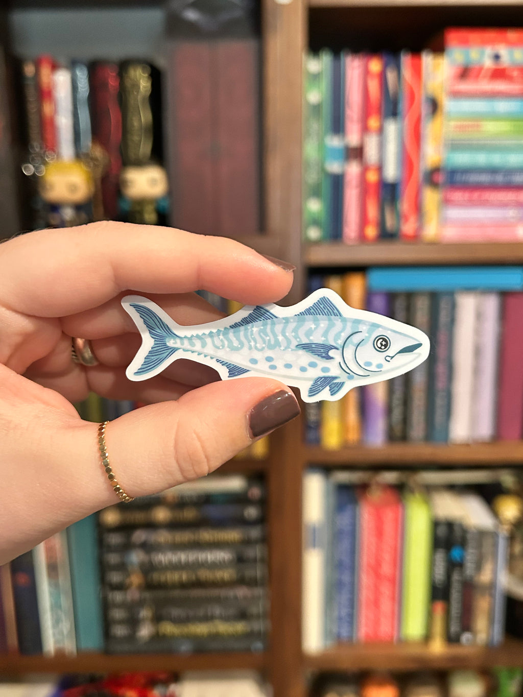 A hand holding a die-cut sticker shaped like a blue and white spotted fish, with a blurred background of bookshelves.