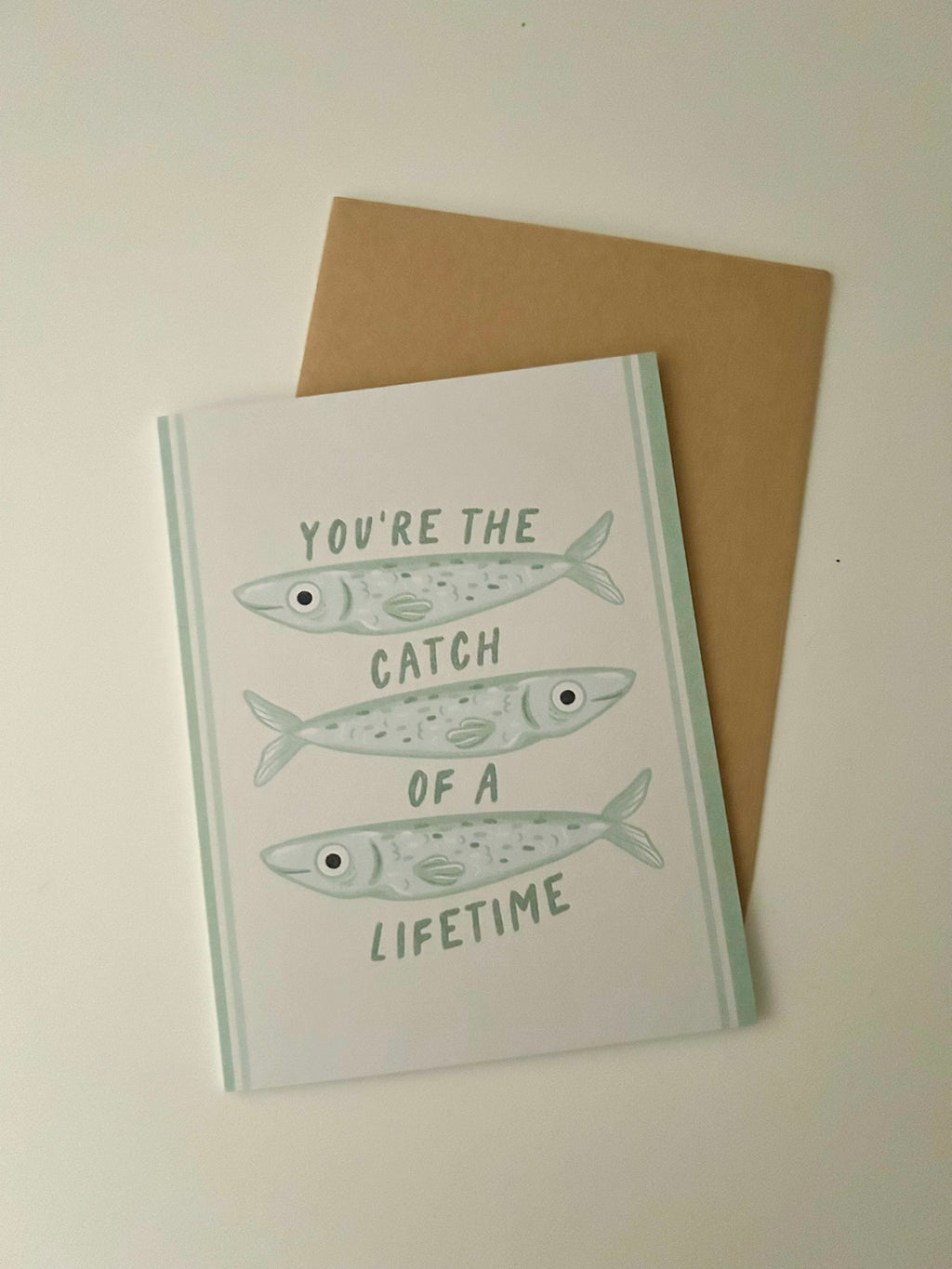 Cute romantic greeting card featuring three illustrated fish and the message "You're the Catch of a Lifetime" on a soft cream background with light blue accents.
