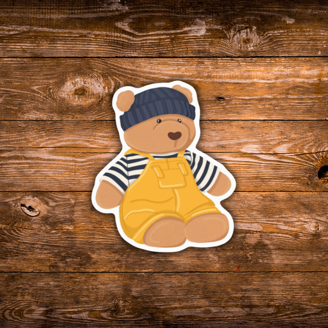 A die-cut sticker of a cartoon teddy bear wearing a navy blue knit hat, a navy and white striped shirt, and yellow overalls, displayed on a rustic brown wooden plank background.
