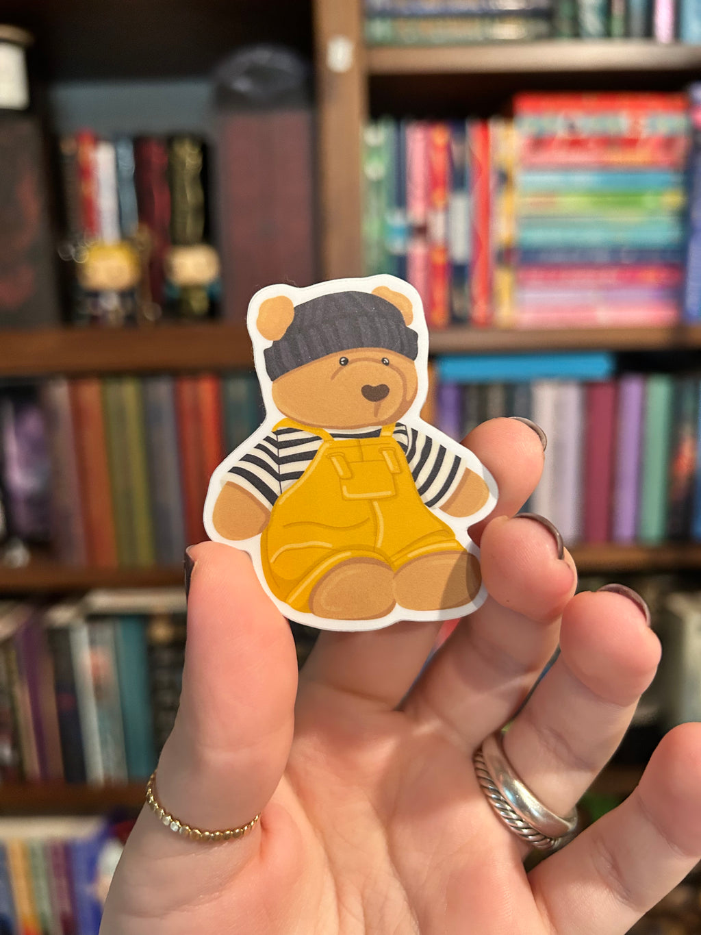 A hand holds a die-cut sticker of a cute teddy bear wearing a dark gray beanie, a black and white striped shirt, and yellow overalls, with a blurred bookshelf in the background.