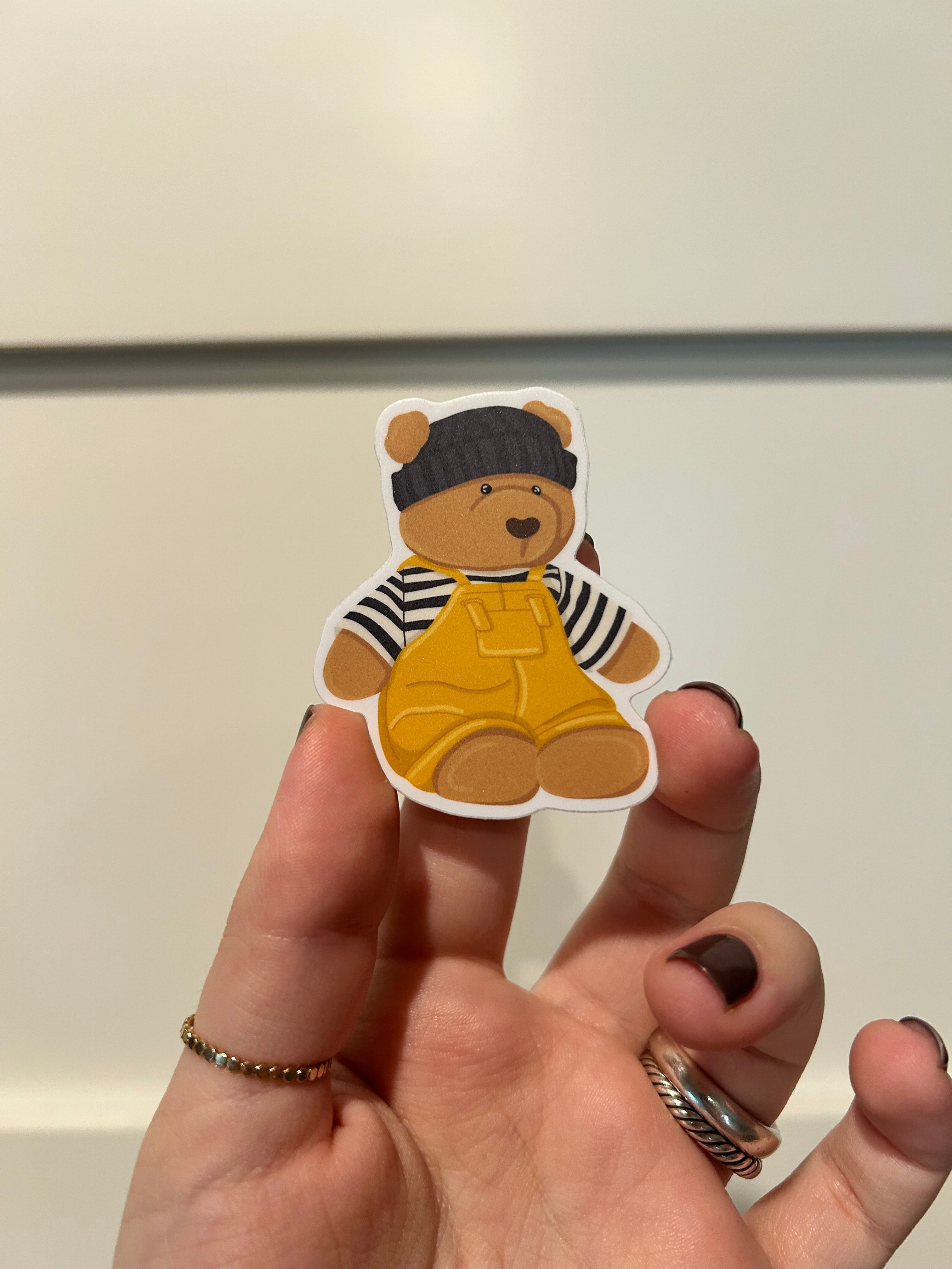A hand with dark nail polish holds a small die-cut sticker of a teddy bear wearing a black-and-white striped shirt and yellow overalls against a plain light background.