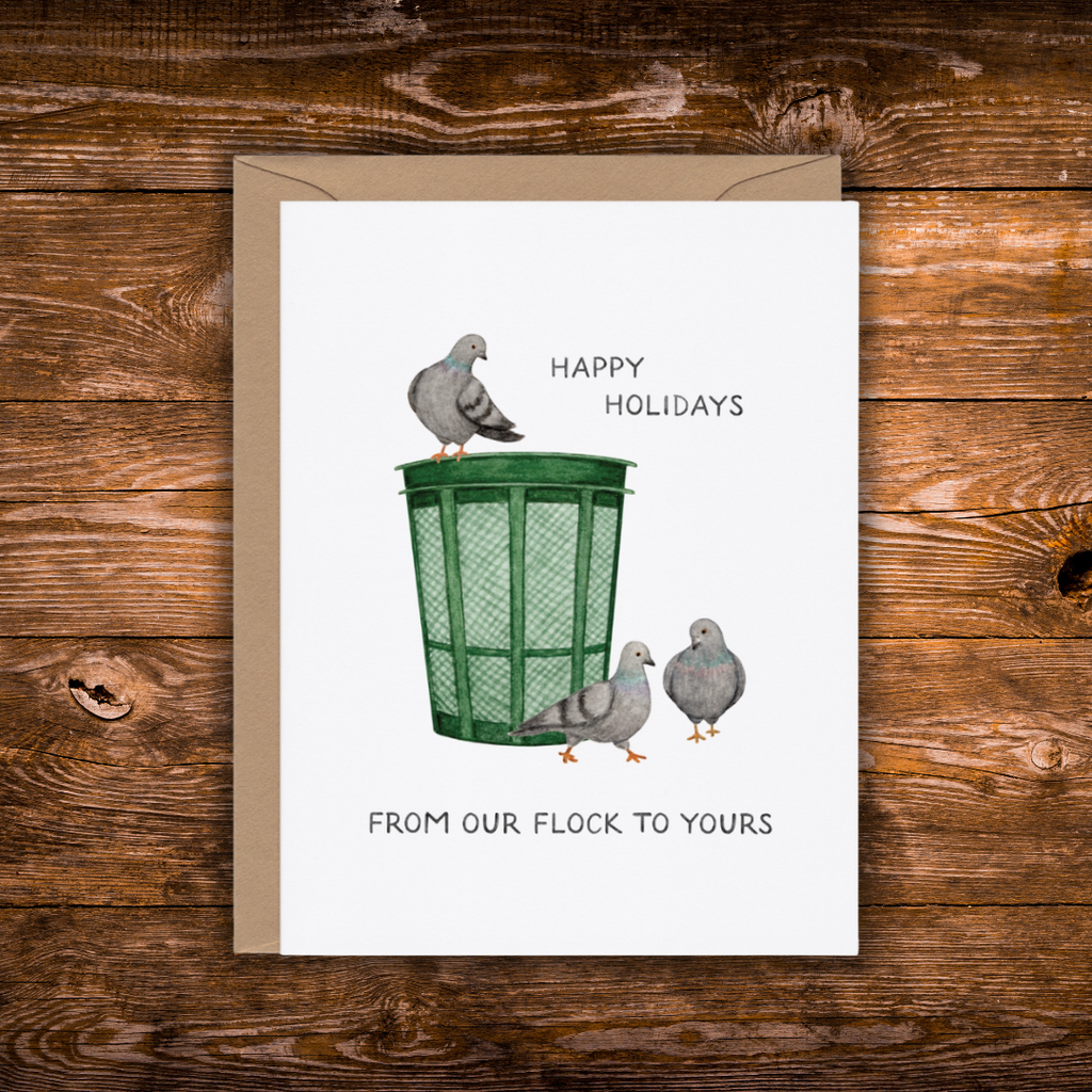 A white greeting card with a brown envelope, featuring a watercolor illustration of three pigeons around a green trash can, with the text 'HAPPY HOLIDAYS' and 'FROM OUR FLOCK TO YOURS', displayed on a rustic wooden background.