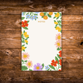 Decorative notepad featuring a colorful floral border with blooming flowers and greenery surrounding a large blank writing area on a cream background.
