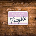 A rectangular sign with rounded corners featuring the text 'BE NICE TO ME I'M Fragile' in white and dark blue lettering on a lavender and cream background, displayed on a rustic brown wooden surface.