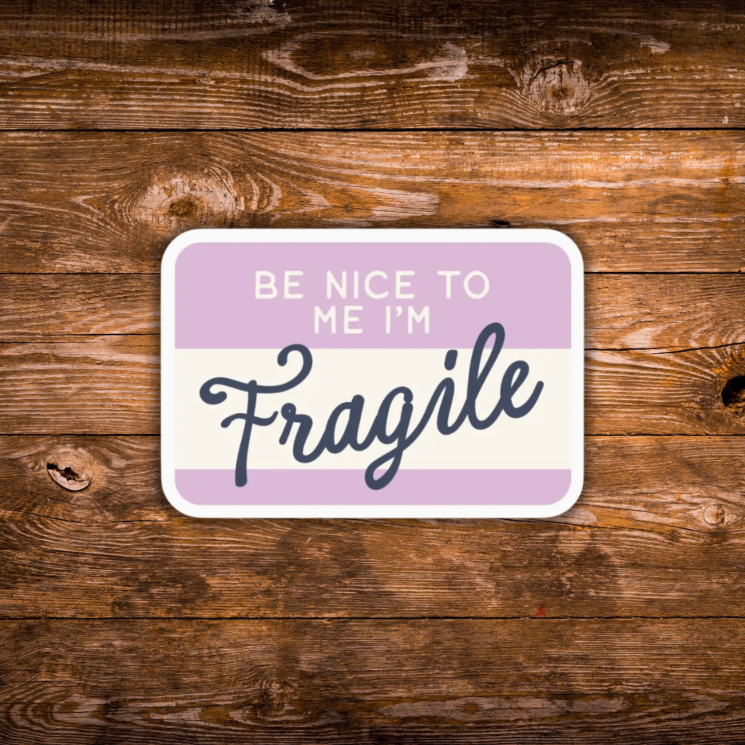 A rectangular sign with rounded corners featuring the text 'BE NICE TO ME I'M Fragile' in white and dark blue lettering on a lavender and cream background, displayed on a rustic brown wooden surface.