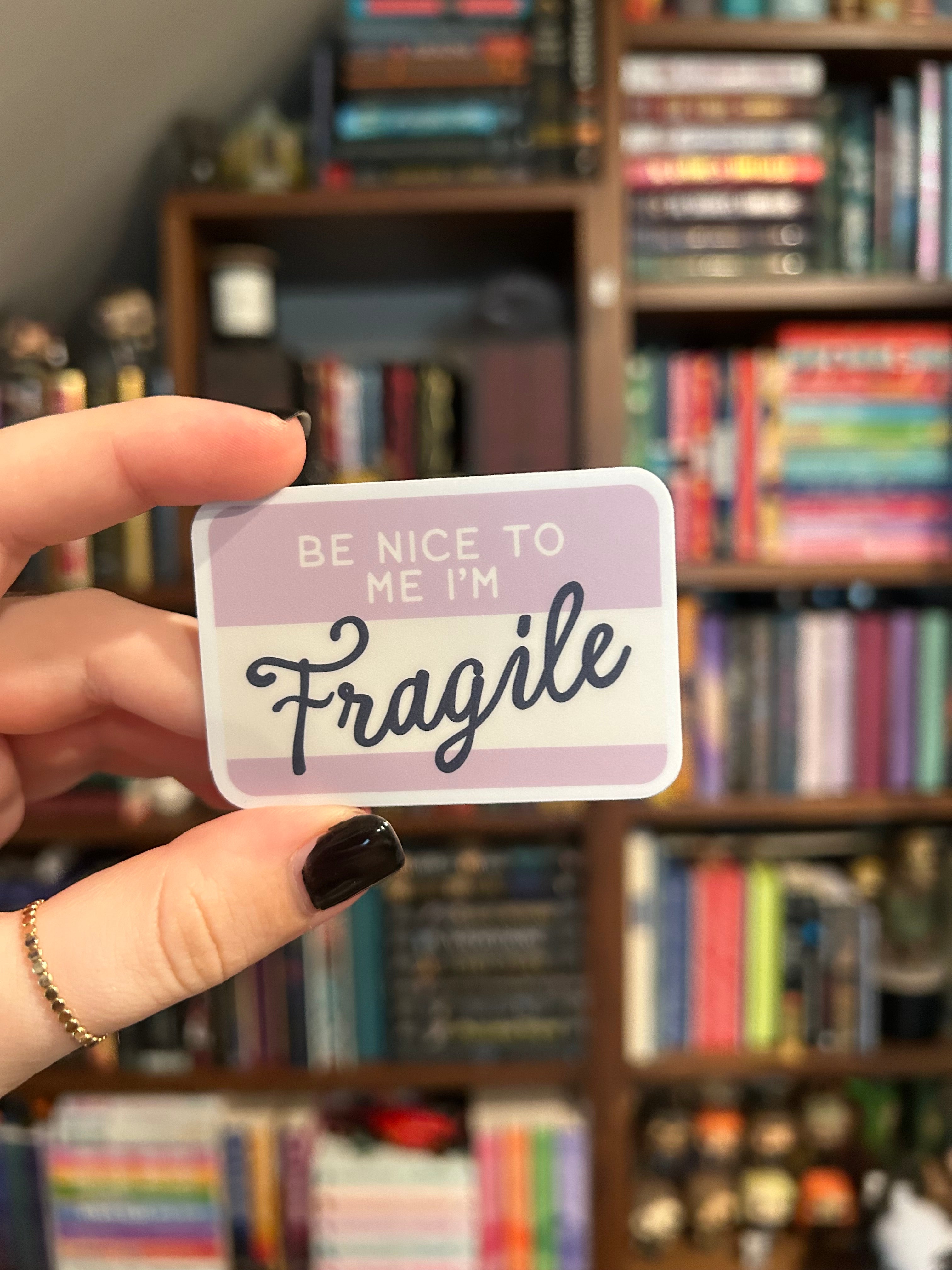 A rectangular card with the words 'BE NICE TO ME I'M Fragile' in black script and block lettering, held by a hand against a blurred background of bookshelves.