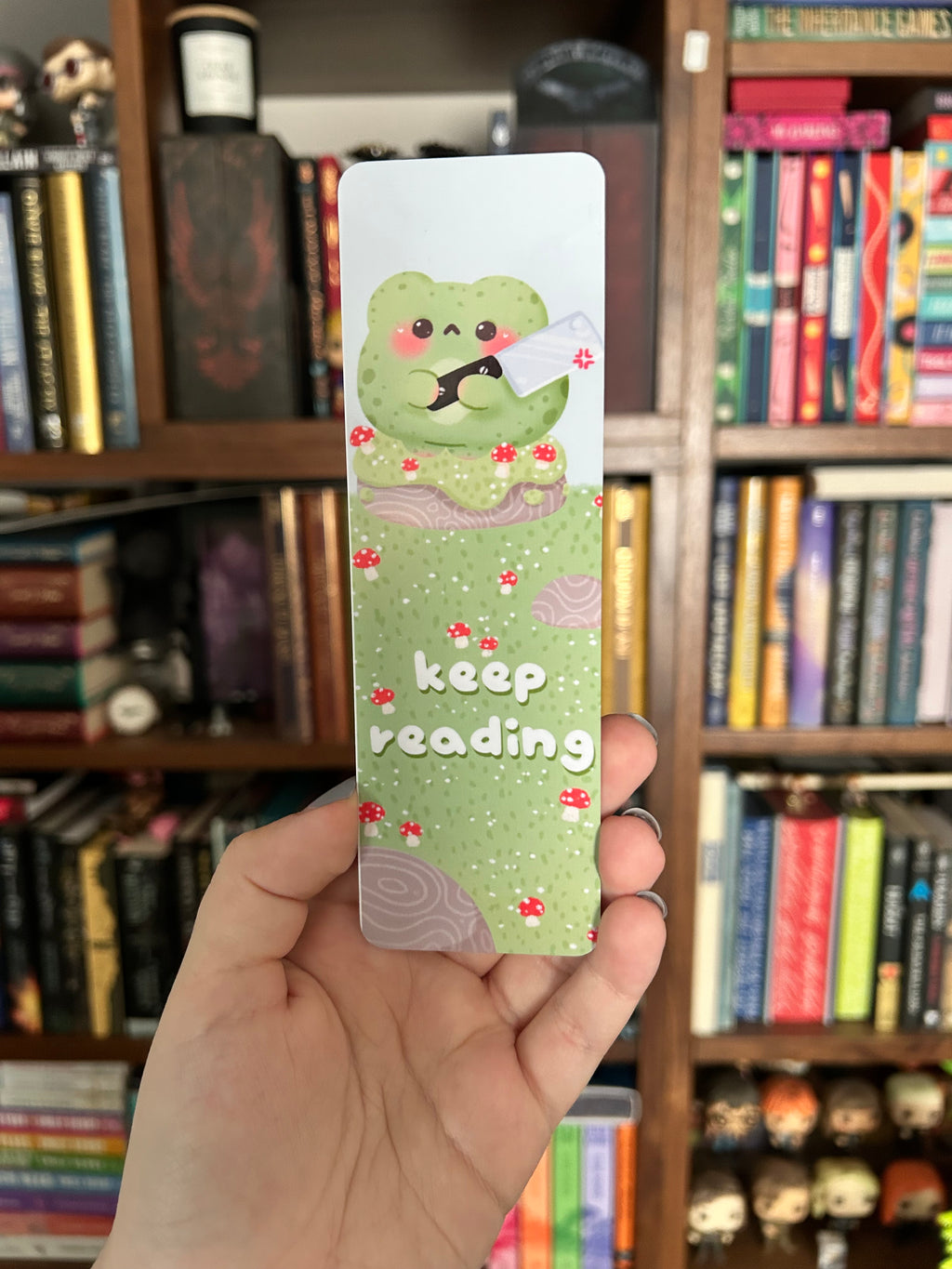 Double-sided frog bookmark featuring a cute green frog holding a cleaver in a mushroom-covered woodland scene with “Keep Reading” lettering, paired with a green checkerboard pattern and pink hearts.