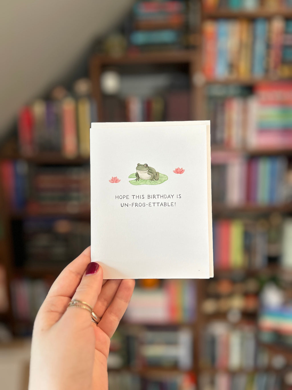 Birthday greeting card featuring a frog sitting on a lily pad with pink water lilies and the phrase "Hope This Birthday Is Un-Frog-Ettable!"