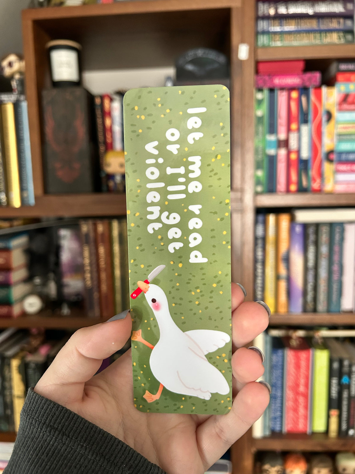 Double-sided goose bookmark featuring a white goose holding a tiny knife and the phrase “Let me read or I will stab” on sage green, with a repeating pattern of geese on the reverse.