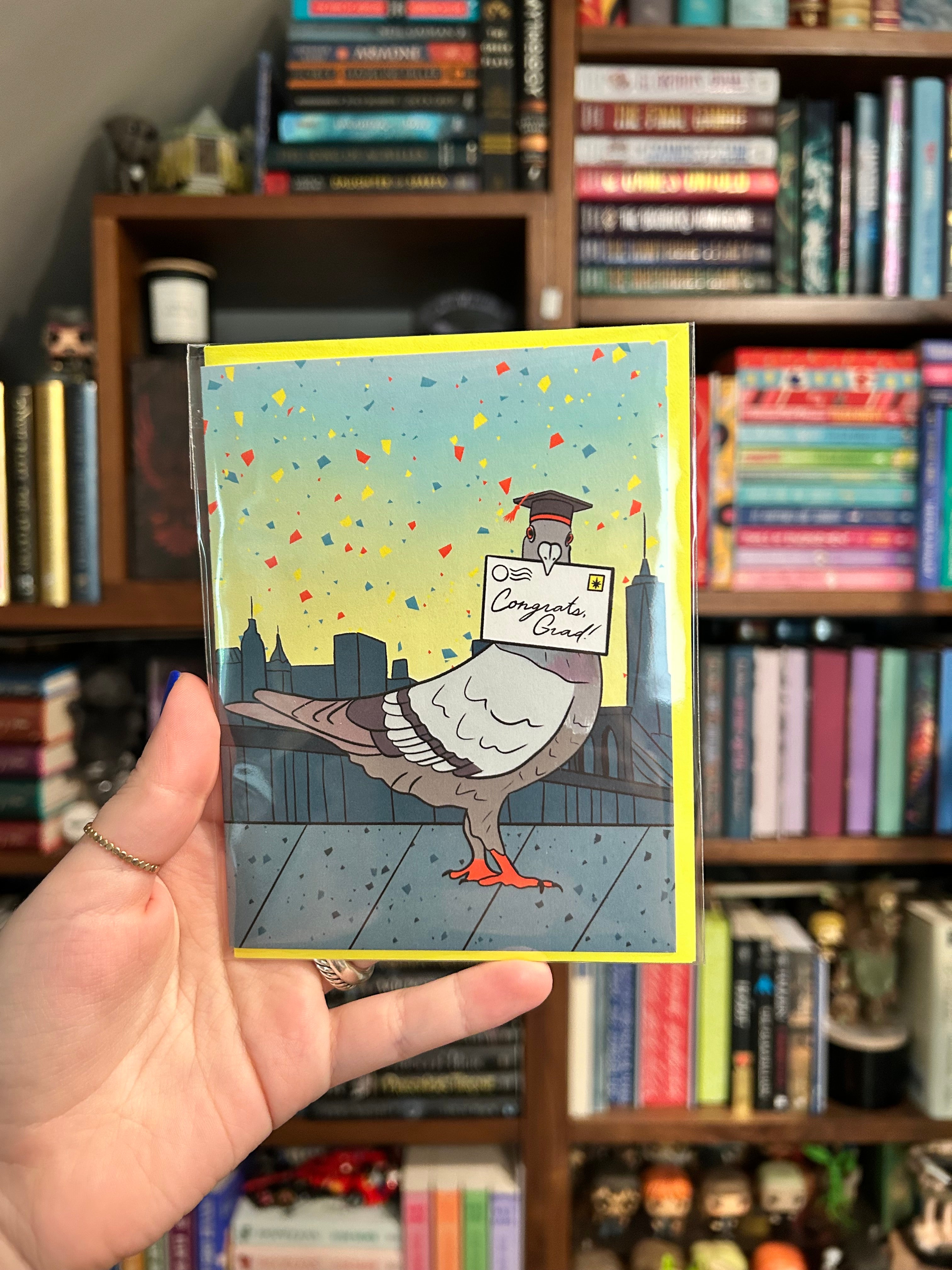 Graduation greeting card featuring a pigeon wearing a graduation cap while delivering a "Congrats, Grad!" letter with the New York City skyline in the background.