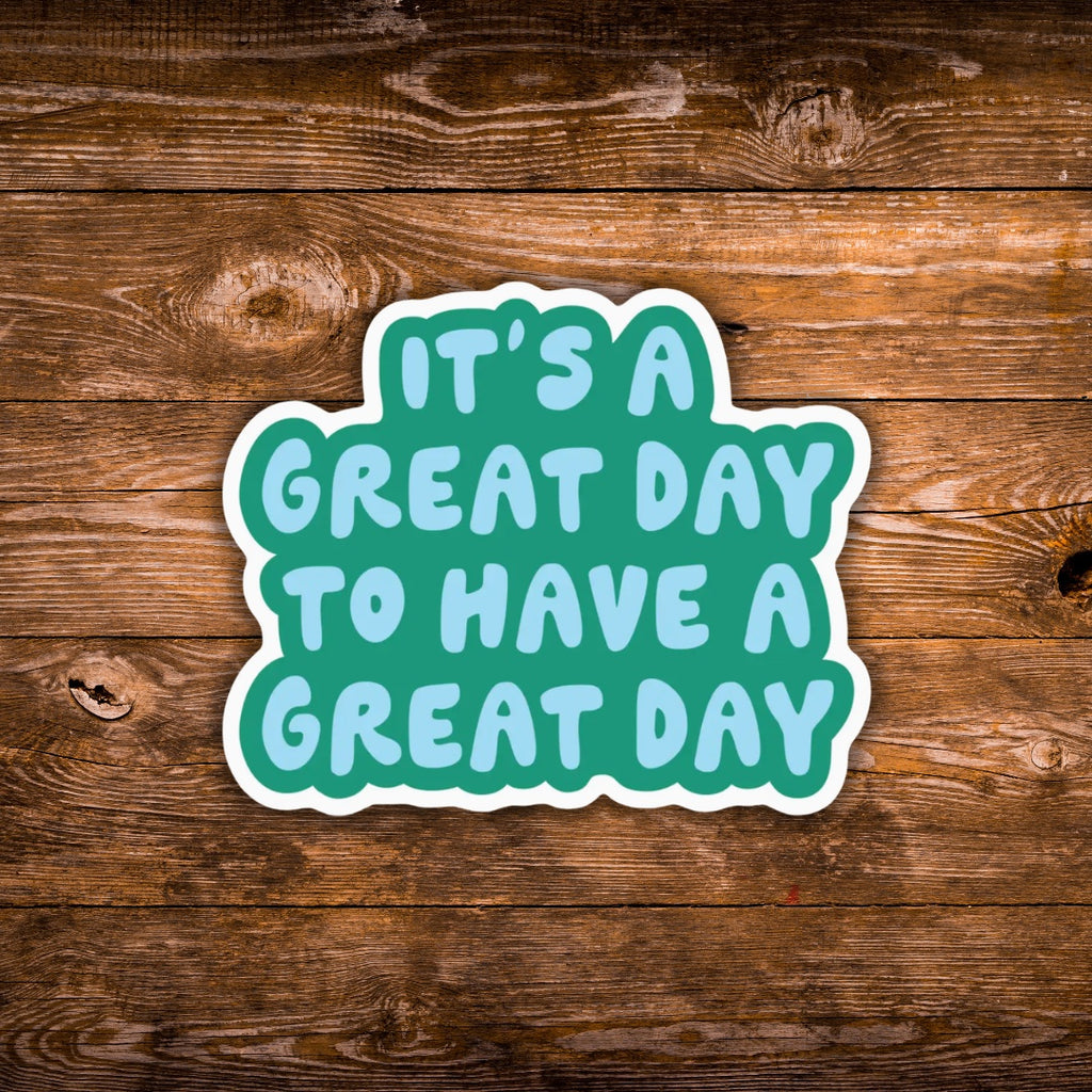 A die-cut vinyl sticker with the text 'IT'S A GREAT DAY TO HAVE A GREAT DAY' in light blue letters on a teal background with a white border, shown on a rustic brown wooden plank surface.