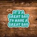 A die-cut vinyl sticker with the text 'IT'S A GREAT DAY TO HAVE A GREAT DAY' in light blue letters on a teal background with a white border, shown on a rustic brown wooden plank surface.