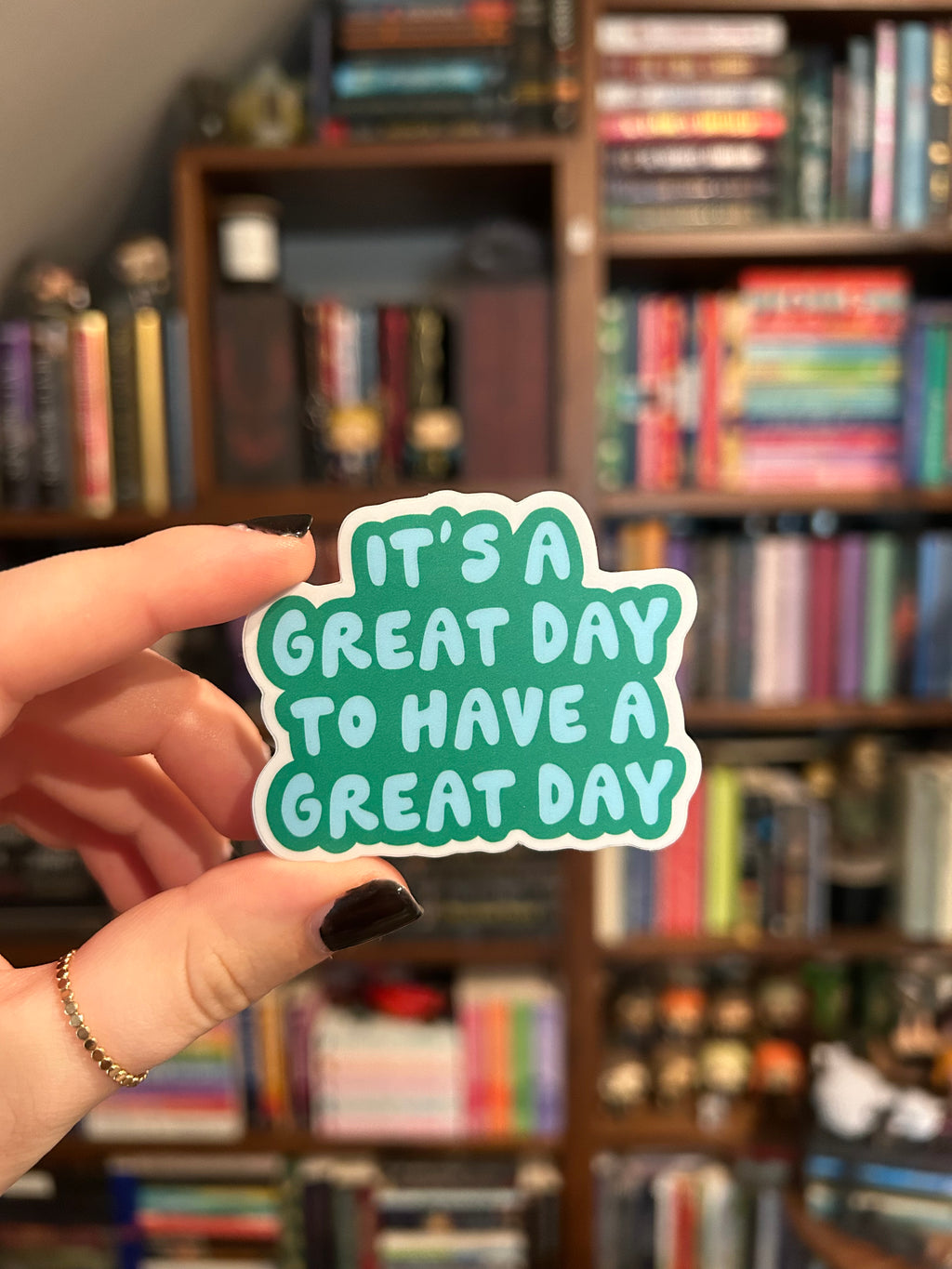 A hand with dark nail polish holds a die-cut sticker that reads 'IT'S A GREAT DAY TO HAVE A GREAT DAY' in white text on a mint green background. The background is a blurred bookshelf.