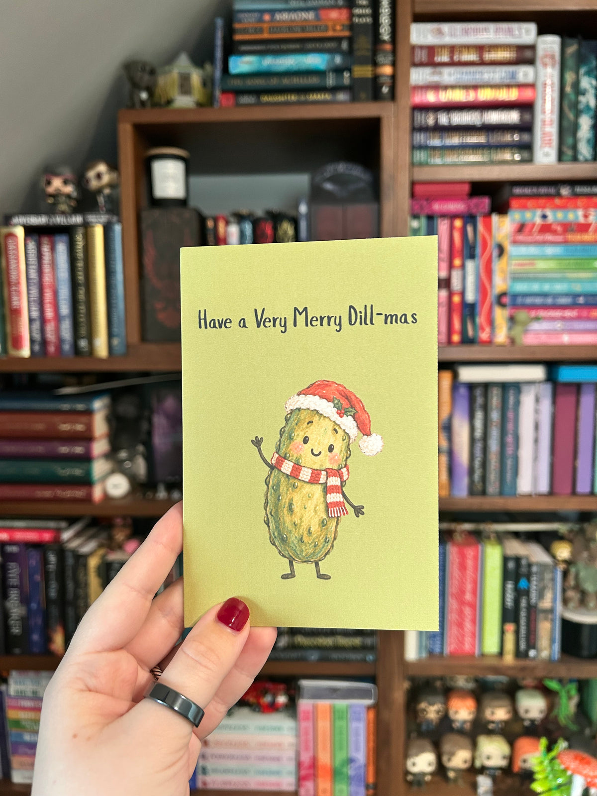 Green Christmas greeting card featuring a smiling green pickle wearing a Santa hat and red-and-white scarf with the phrase “Have a Very Merry Dill-mas.” 