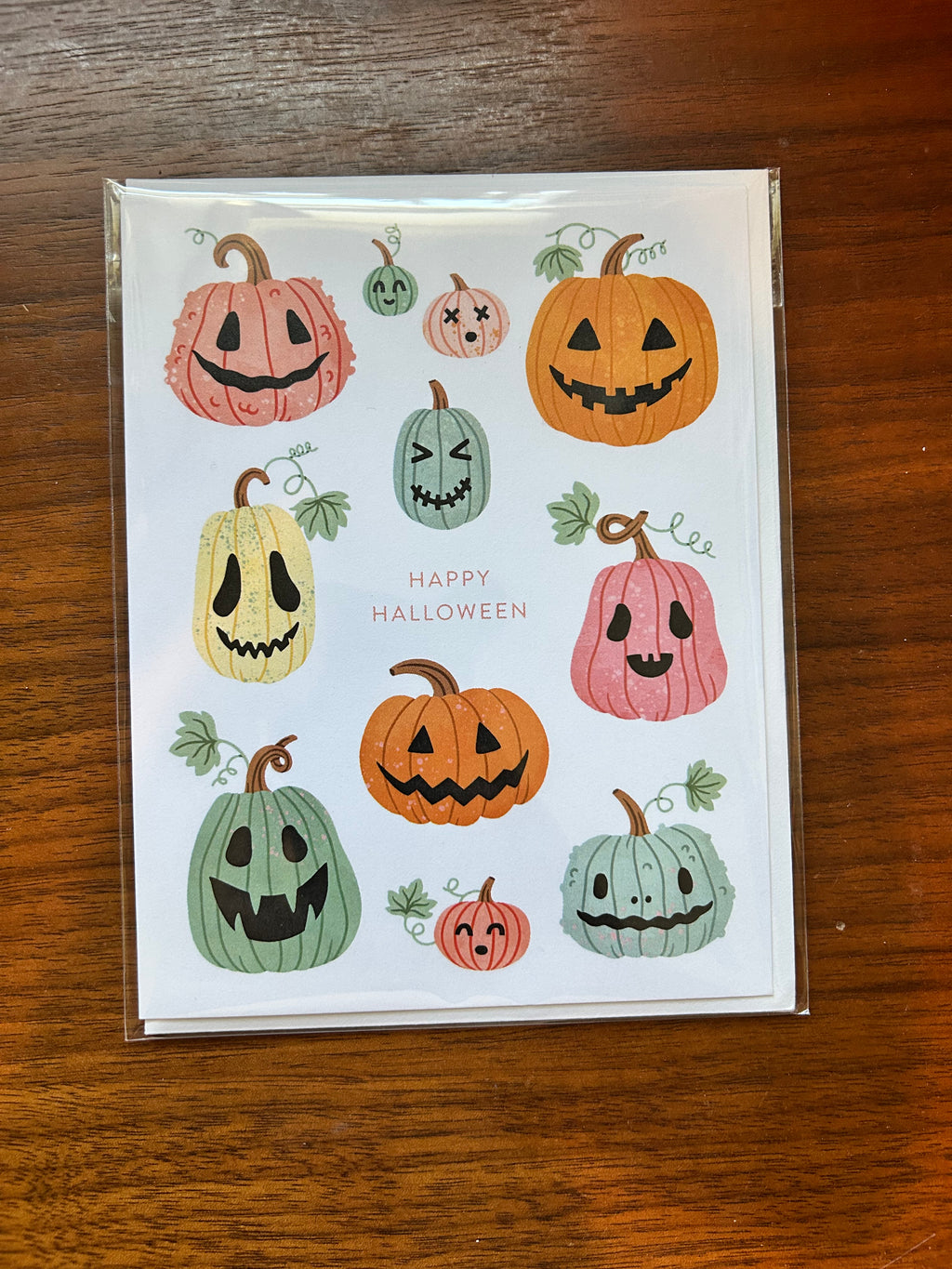 A square white greeting card featuring a collection of hand-drawn, colorful jack-o'-lantern pumpkins in orange, pink, yellow, and green, with the words 'Happy Halloween' printed vertically. The card is enclosed in a clear plastic sleeve and placed on a dark brown wooden surface.