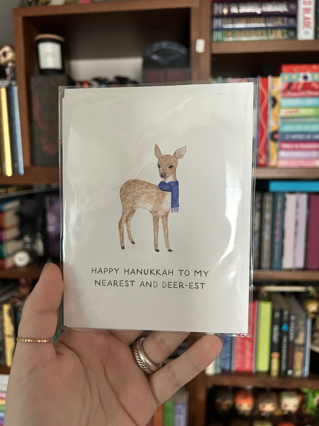 A hand holding a Hanukkah greeting card with a watercolor illustration of a deer wearing a blue sweater. The card reads 'HAPPY HANUKKAH TO MY NEAREST AND DEER-EST' and is photographed in front of a blurred bookshelf background.