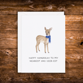 A white greeting card with a watercolor deer wearing a blue scarf and the text 'HAPPY HANUKKAH TO MY NEAREST AND DEER-EST', placed on top of a white envelope against a rustic brown wooden background.