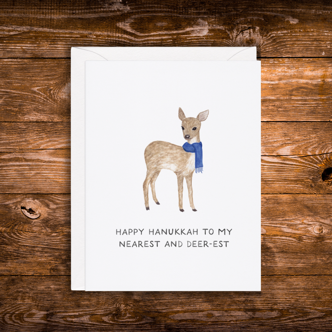 A white greeting card with a watercolor deer wearing a blue scarf and the text 'HAPPY HANUKKAH TO MY NEAREST AND DEER-EST', placed on top of a white envelope against a rustic brown wooden background.