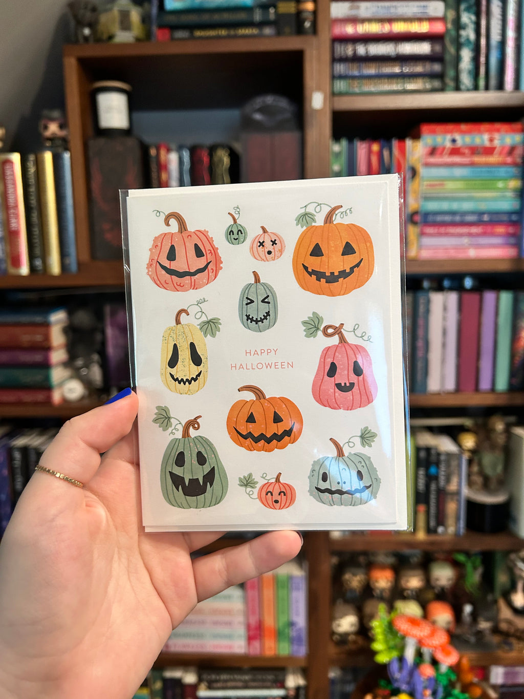 A hand holds a printed card featuring illustrated jack-o'-lantern pumpkins in orange, pink, yellow, and green on a white background, with the words 'HAPPY HALLOWEEN'. The background shows a blurred bookstore or library shelf filled with books.