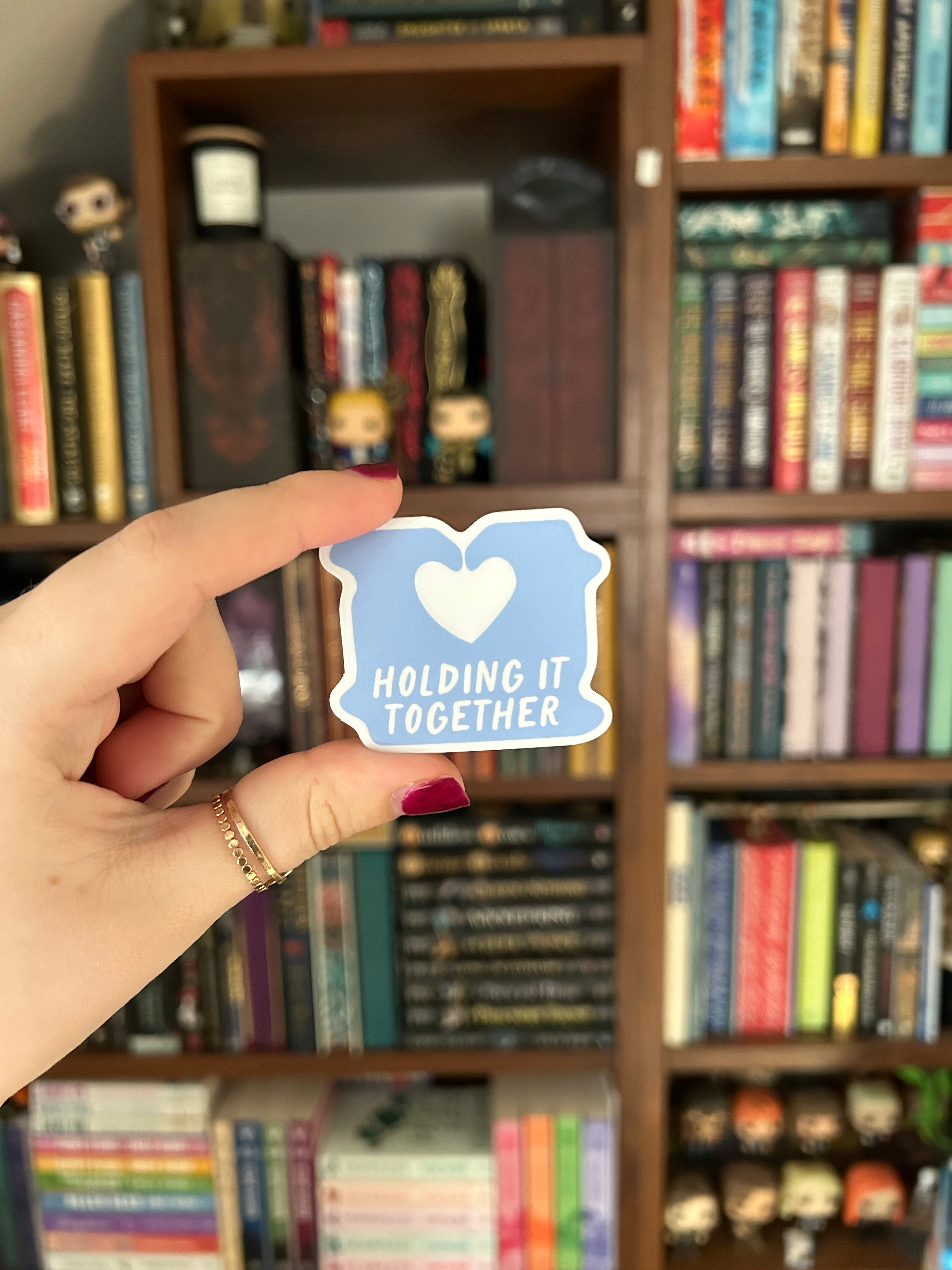 A hand with red nail polish holds a small blue sticker that says 'HOLDING IT TOGETHER' with a white heart, in front of a blurred bookshelf background.