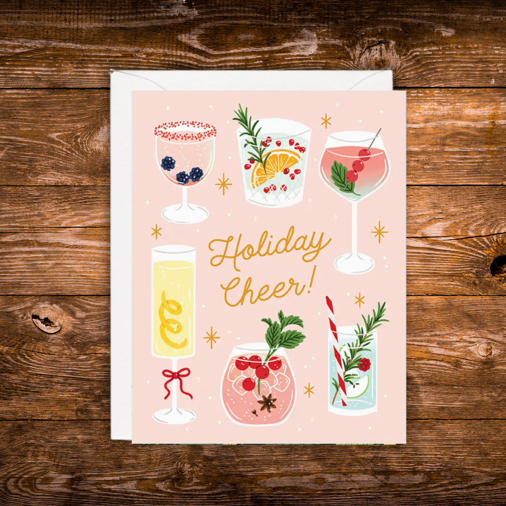A square greeting card with a white envelope, featuring a pastel pink background and illustrated holiday drinks, with the text 'Holiday Cheer!' in gold script. The card is photographed flat on a rustic dark wood surface.