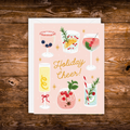 A square greeting card with a white envelope, featuring a pastel pink background and illustrated holiday drinks, with the text 'Holiday Cheer!' in gold script. The card is photographed flat on a rustic dark wood surface.
