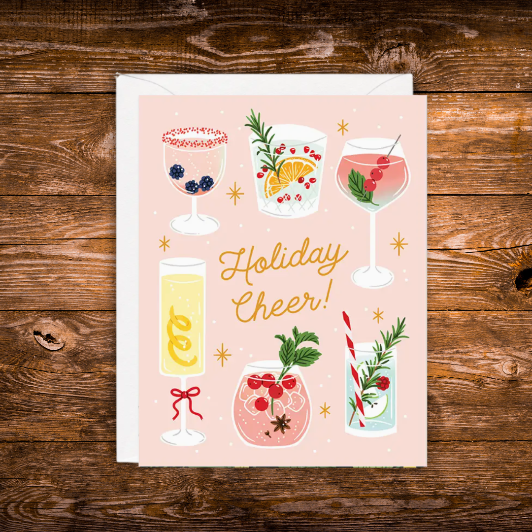 A square greeting card with a white envelope, featuring a pastel pink background and illustrated holiday drinks, with the text 'Holiday Cheer!' in gold script. The card is photographed flat on a rustic dark wood surface.