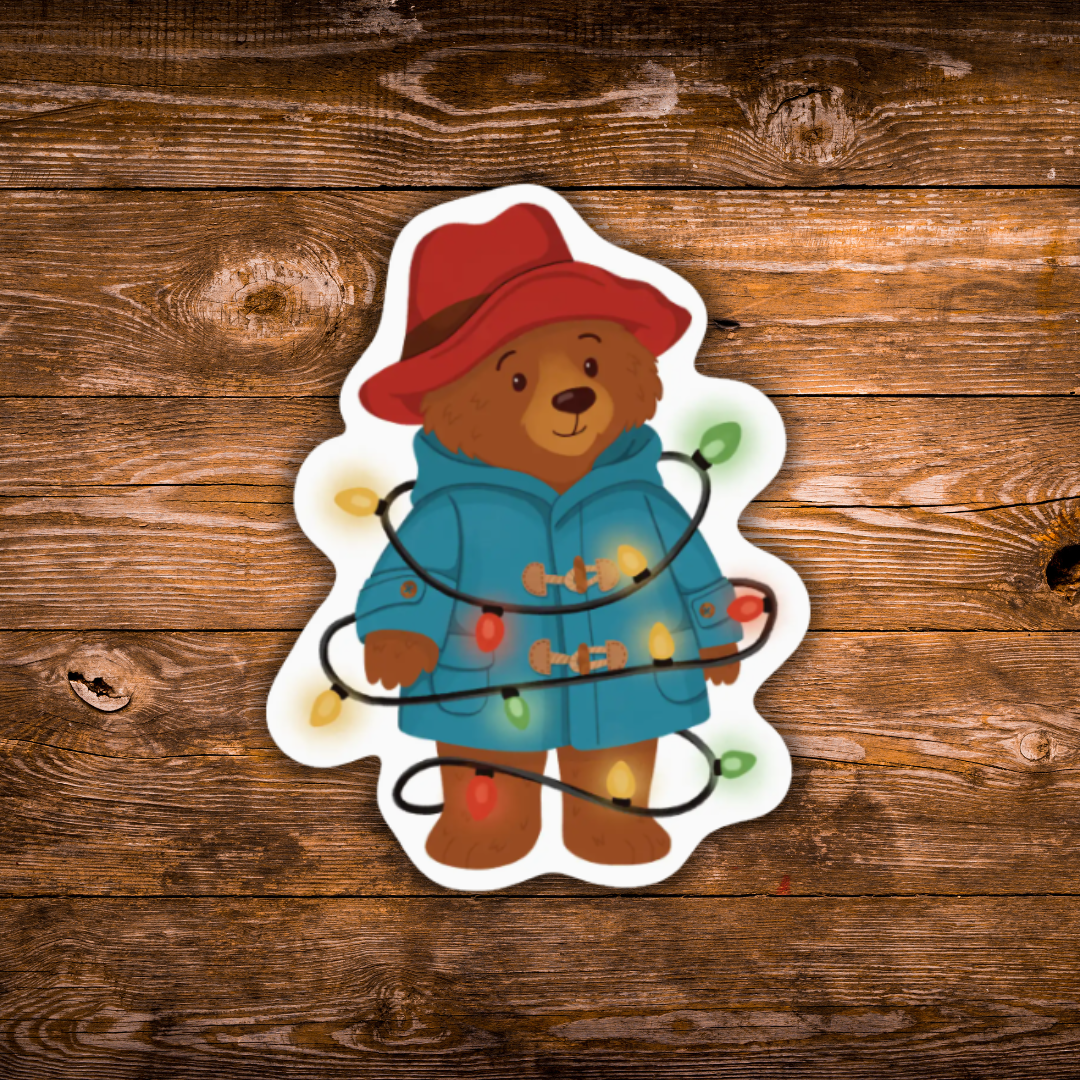 A die-cut sticker featuring an illustrated brown bear wearing a red hat and blue coat wrapped in holiday lights, shown on a rustic brown wood plank background.