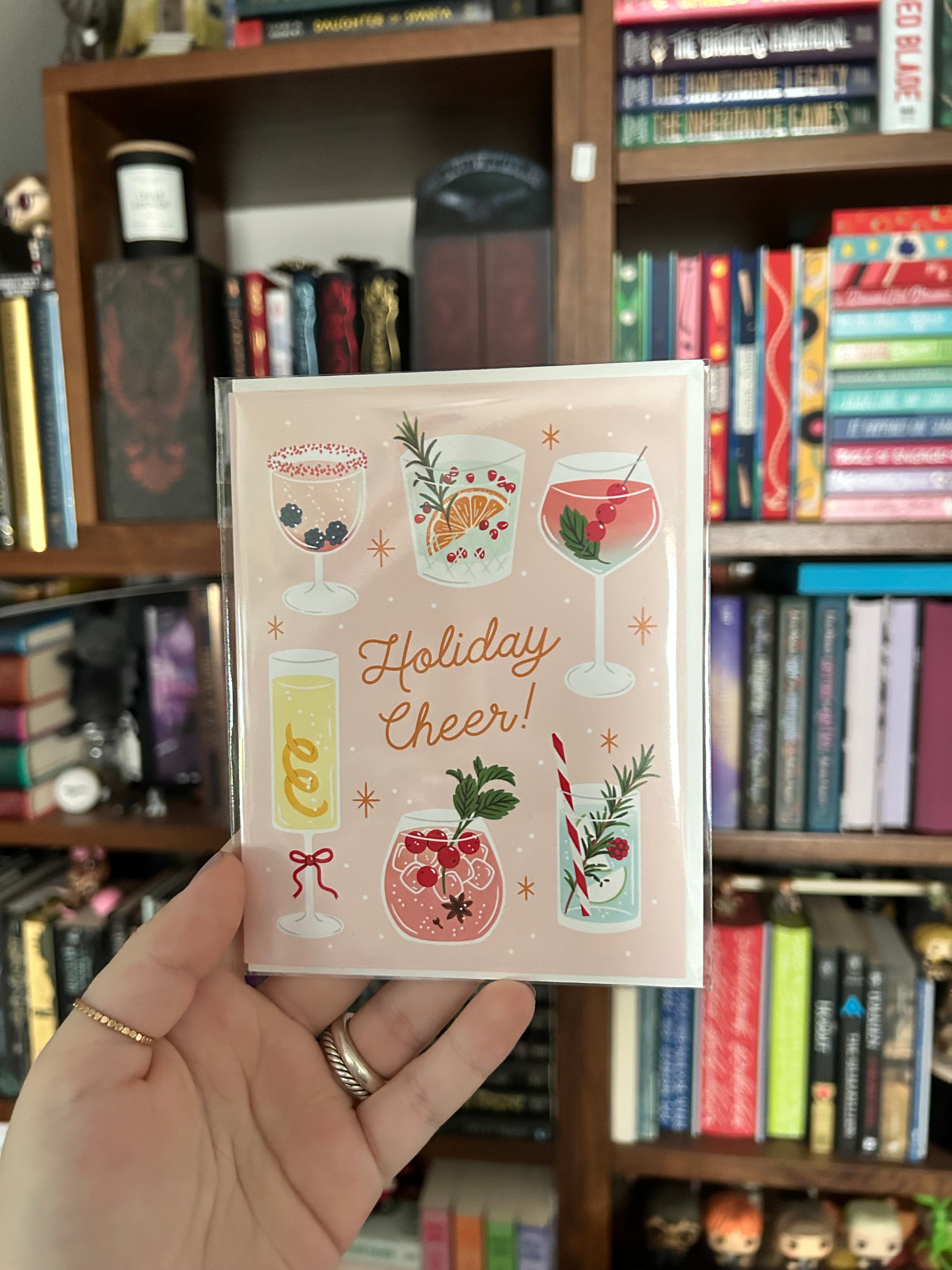 A hand holding a square greeting card that reads 'Holiday Cheer!' with illustrated holiday drinks, set against a background of wooden bookshelves filled with books.