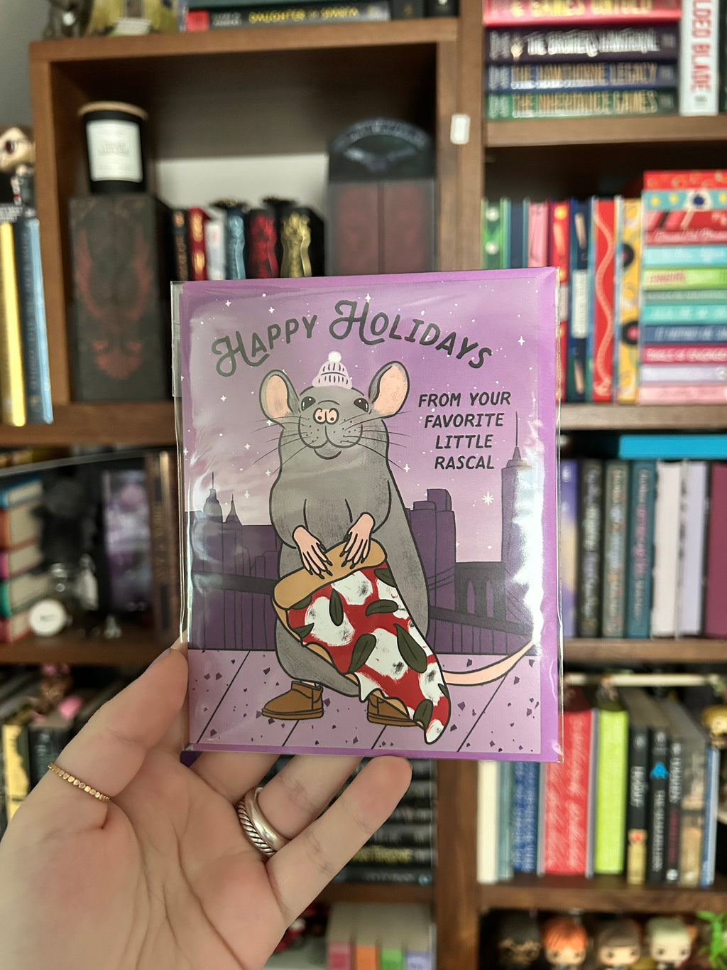 A hand holds a holiday greeting card in front of a bookshelf. The card has a lavender background and features an illustration of a gray rat wearing a white hat and boots, wrapped in a red, white, and black patterned blanket.