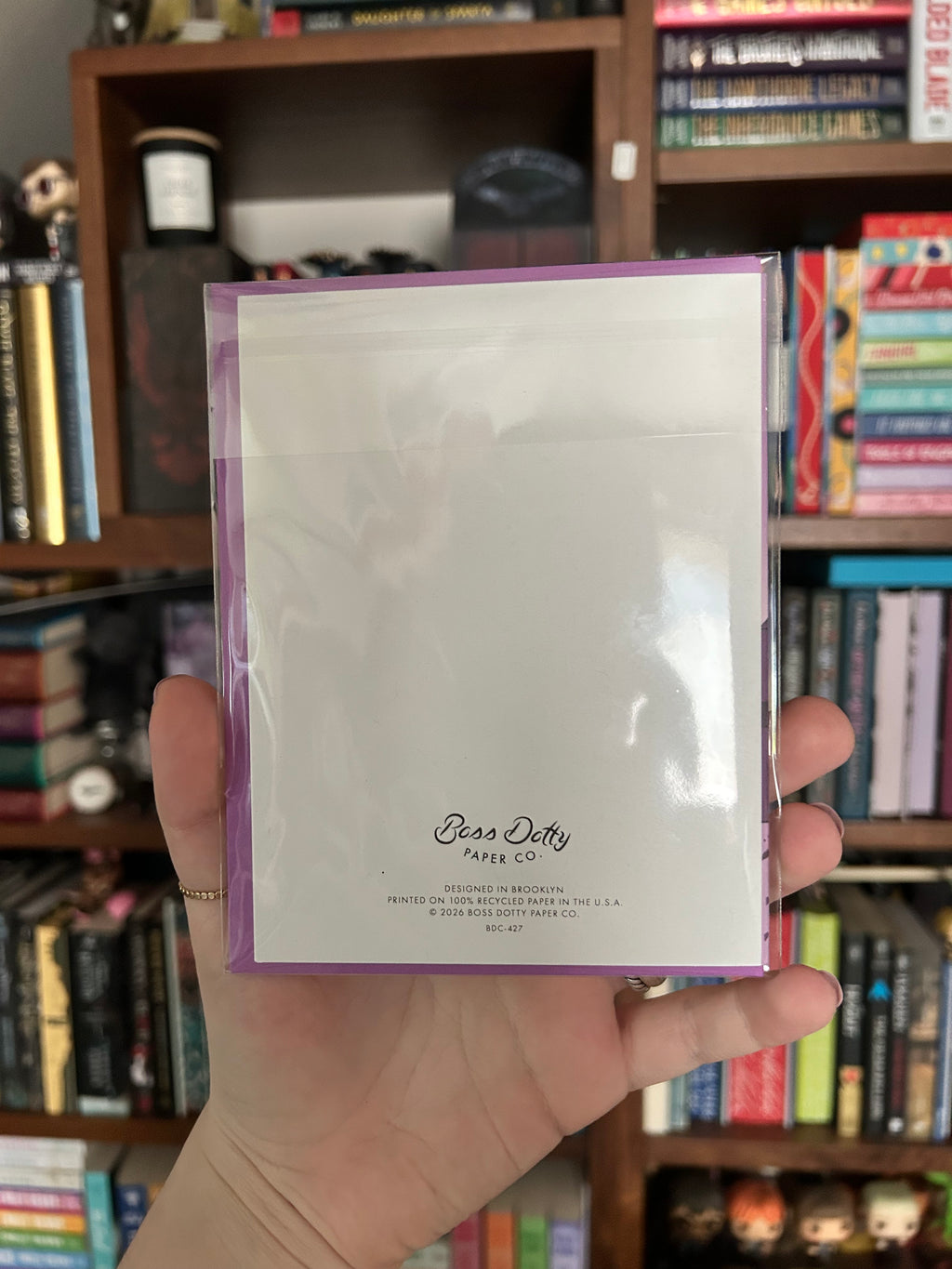 A hand holding up the back of a Christmas card with the brand name 'Boss Dotty Paper Co.' printed near the bottom, set against a blurred bookshelf background.