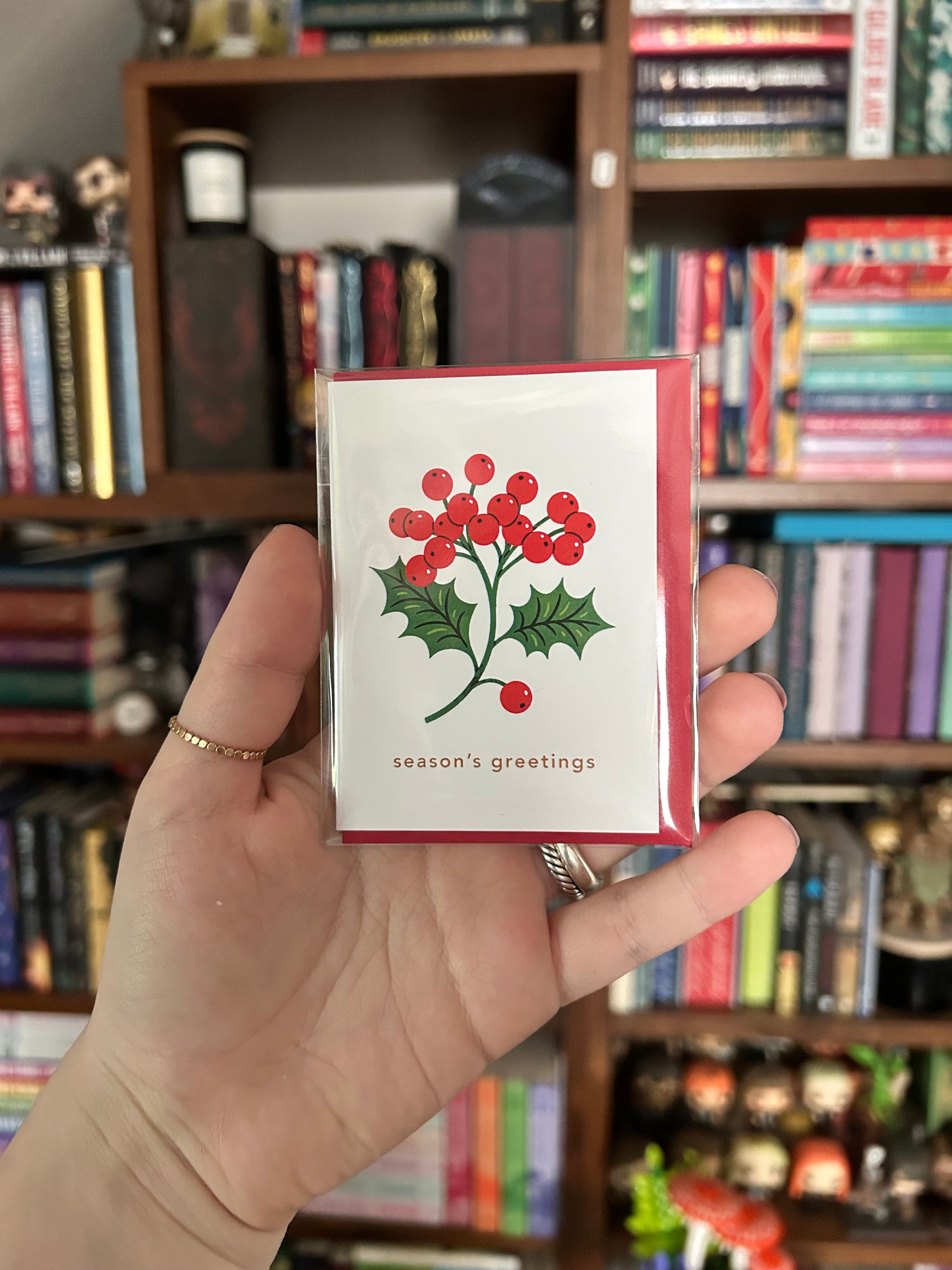 A hand holding a small holiday greeting card with a red border and a holly illustration featuring red berries and green leaves, with the text 'season's greetings' below. The background shows a blurred bookshelf filled with books.